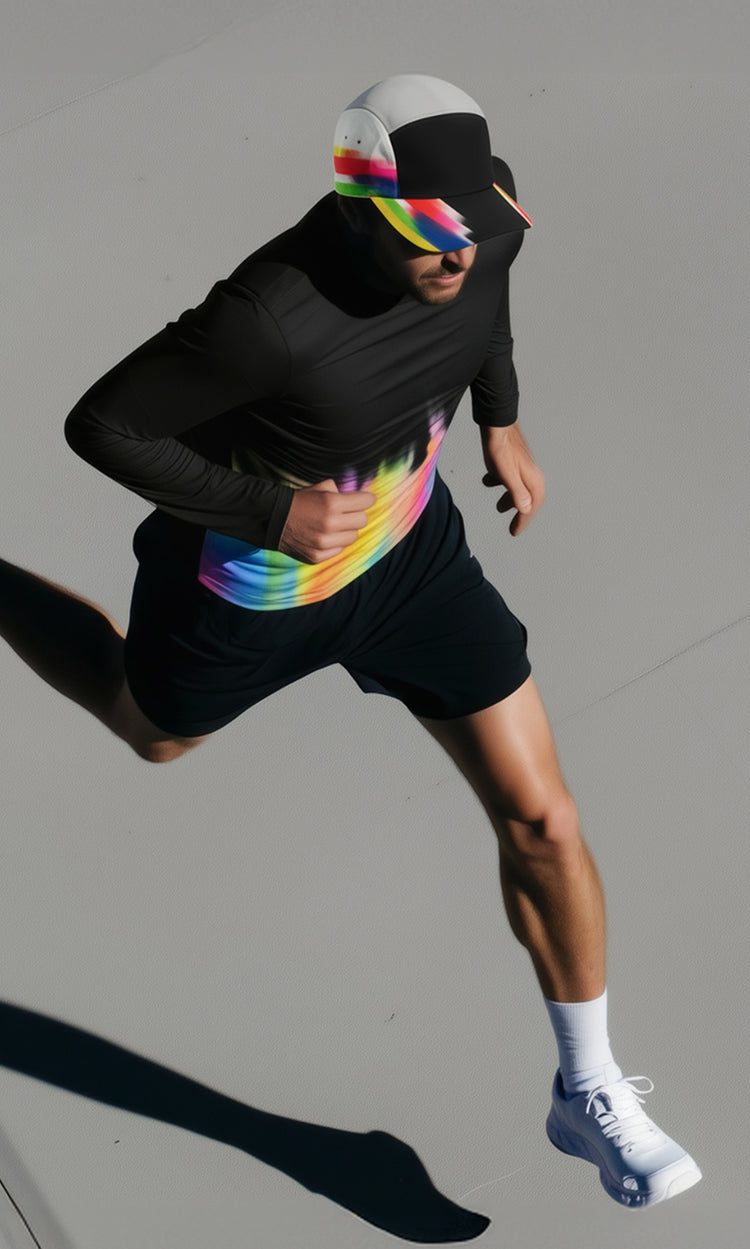 An overhead view of a runner in motion wearing a black long-sleeve top with a rainbow gradient band across the torso, black shorts, white sneakers, and a white cap with rainbow accents, casting a sharp shadow on a light pavement surface.