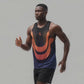muscular Black man wearing a black and orange athletic tank top and black shorts is running against a light neutral background, with his right arm bent forward mid-stride and a focused expression on his face.