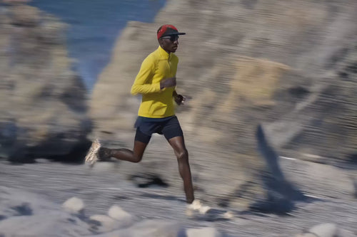 A runner wearing a bright yellow long-sleeve top, navy shorts, a red cap, and sunglasses sprints along a rocky coastal trail, with blurred cliffs and blue water in the background conveying motion.