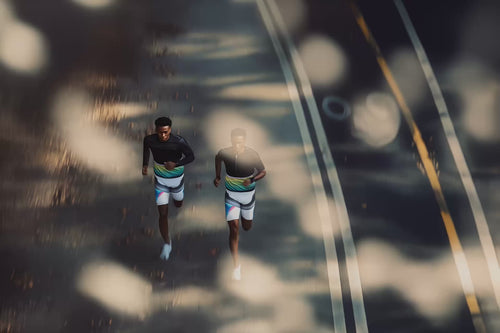 An overhead view of two runners moving side by side on a sun-dappled road, both wearing coordinated athletic outfits with dark tops and patterned shorts, as long shadows and blurred light spots create a dynamic sense of motion.