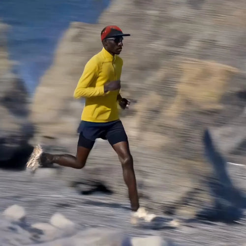A runner wearing a bright yellow long-sleeve top, navy shorts, a red cap, and sunglasses sprints along a rocky coastal trail, with blurred cliffs and blue water in the background conveying motion.